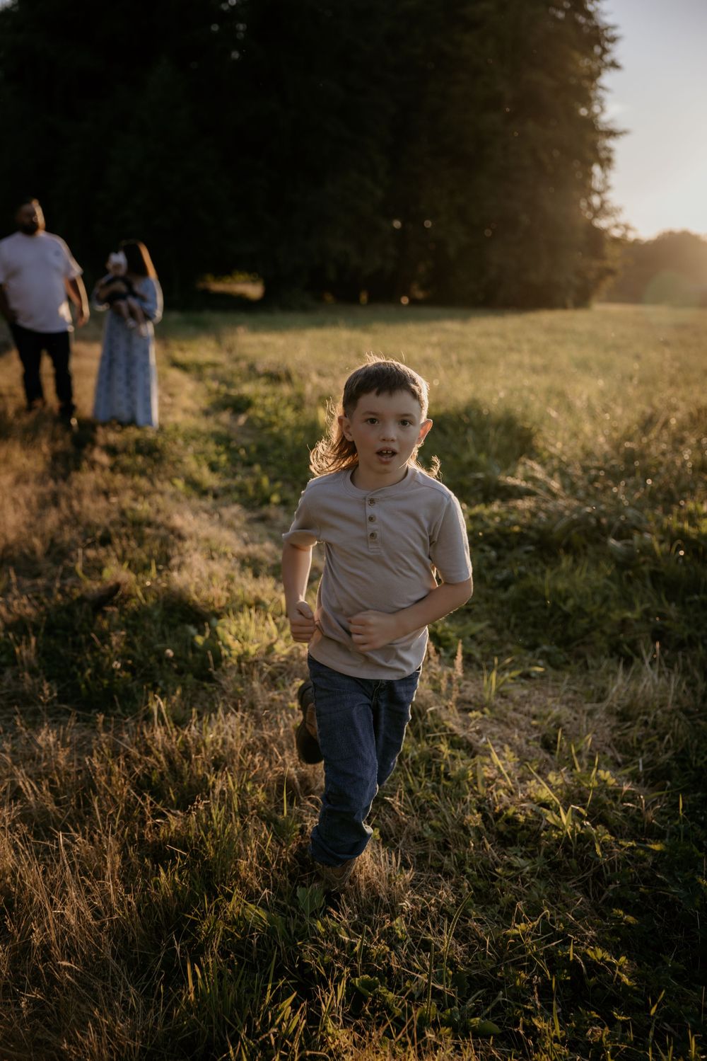 A young boy runs through a sunlit grassy field at golden hour with family standing in the background.
