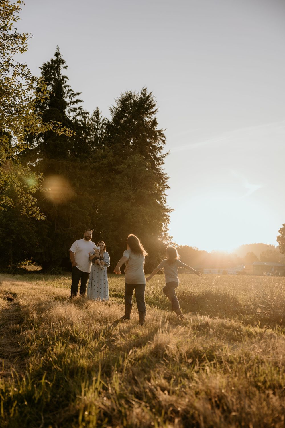 Family walking through golden sunlit meadow at sunset with tall evergreen trees in background.