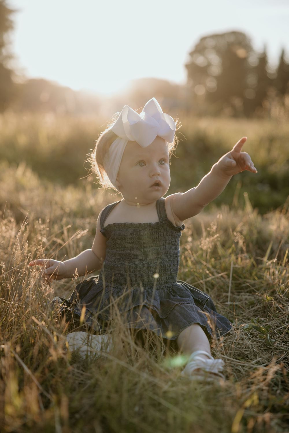 Toddler girl in green dress and white bow sits in golden field, pointing at something during sunset.