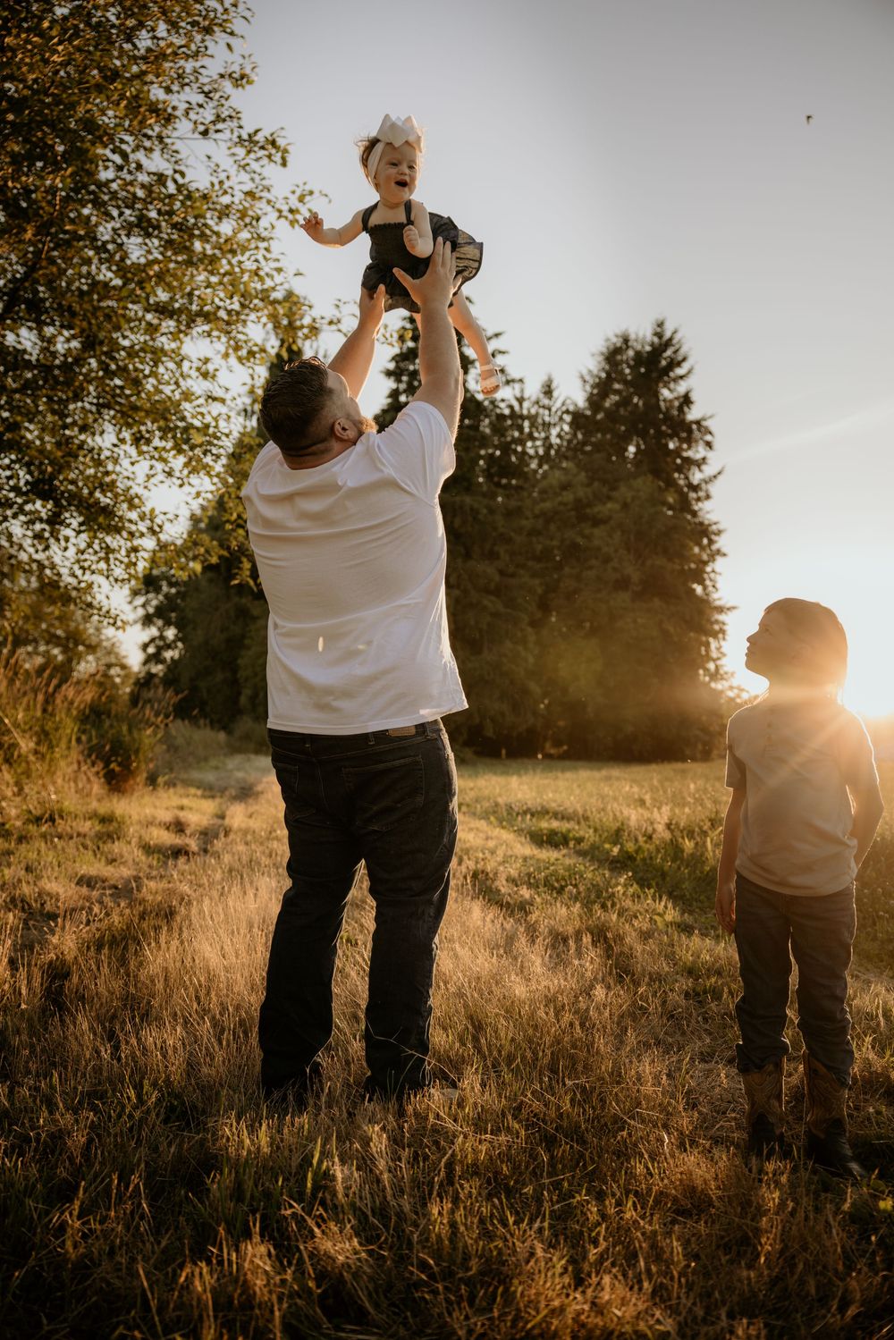 Father tossing laughing child in the air at golden hour in a meadow, with another child watching nearby.