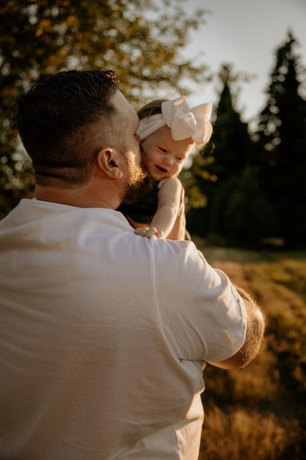 A father holds his smiling baby girl wearing a white bow headband outdoors in warm golden hour light.