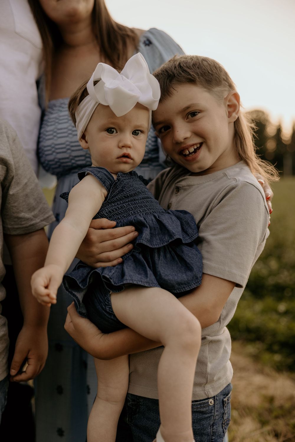 Older girl smiling while holding baby sister in blue dress with white bow outdoors at golden hour.