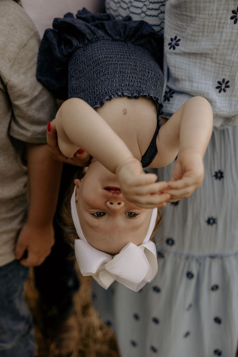 A young child held upside down by parents, wearing a white bow headband and blue dress, laughing joyfully.