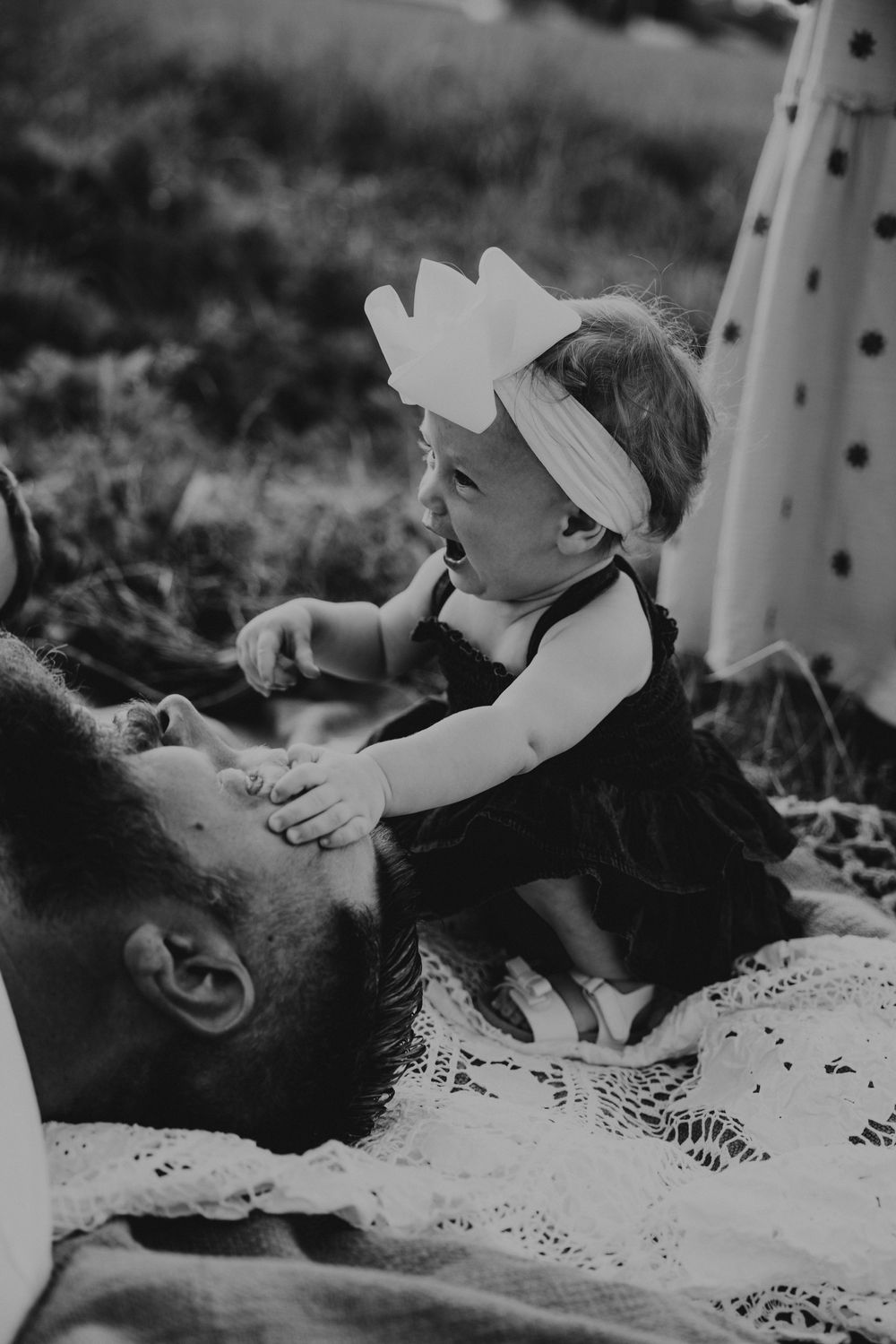 Black and white photo of a bearded man lying down while a smiling baby girl in a bow headband plays on his chest.