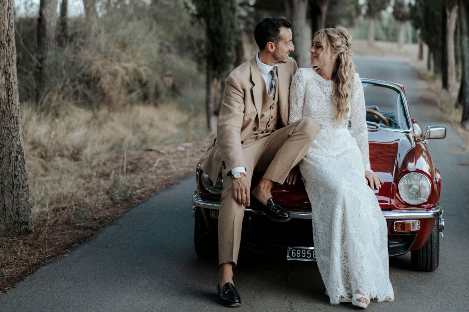 Bride and groom posing together leaning on a vintage red classic car on a tree-lined road on their wedding day.