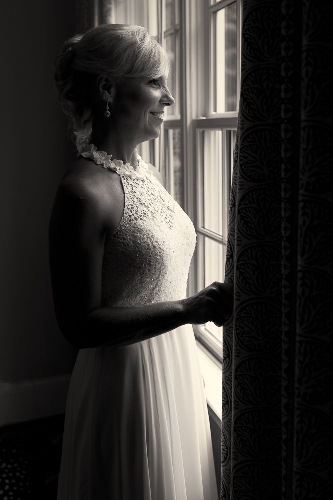 Bride in white halter wedding gown adjusting her heels while seated by a window in a sepia-toned photo.