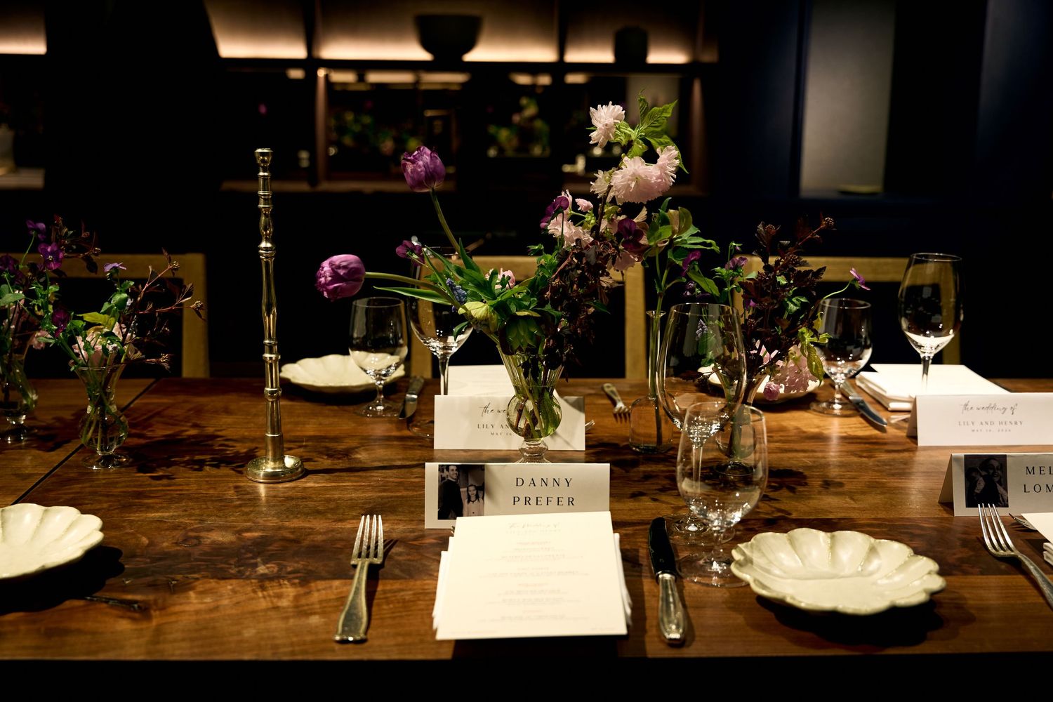 Elegant rustic wooden dining table set with floral centerpieces, candles, wine glasses, and place cards for a formal dinner event.