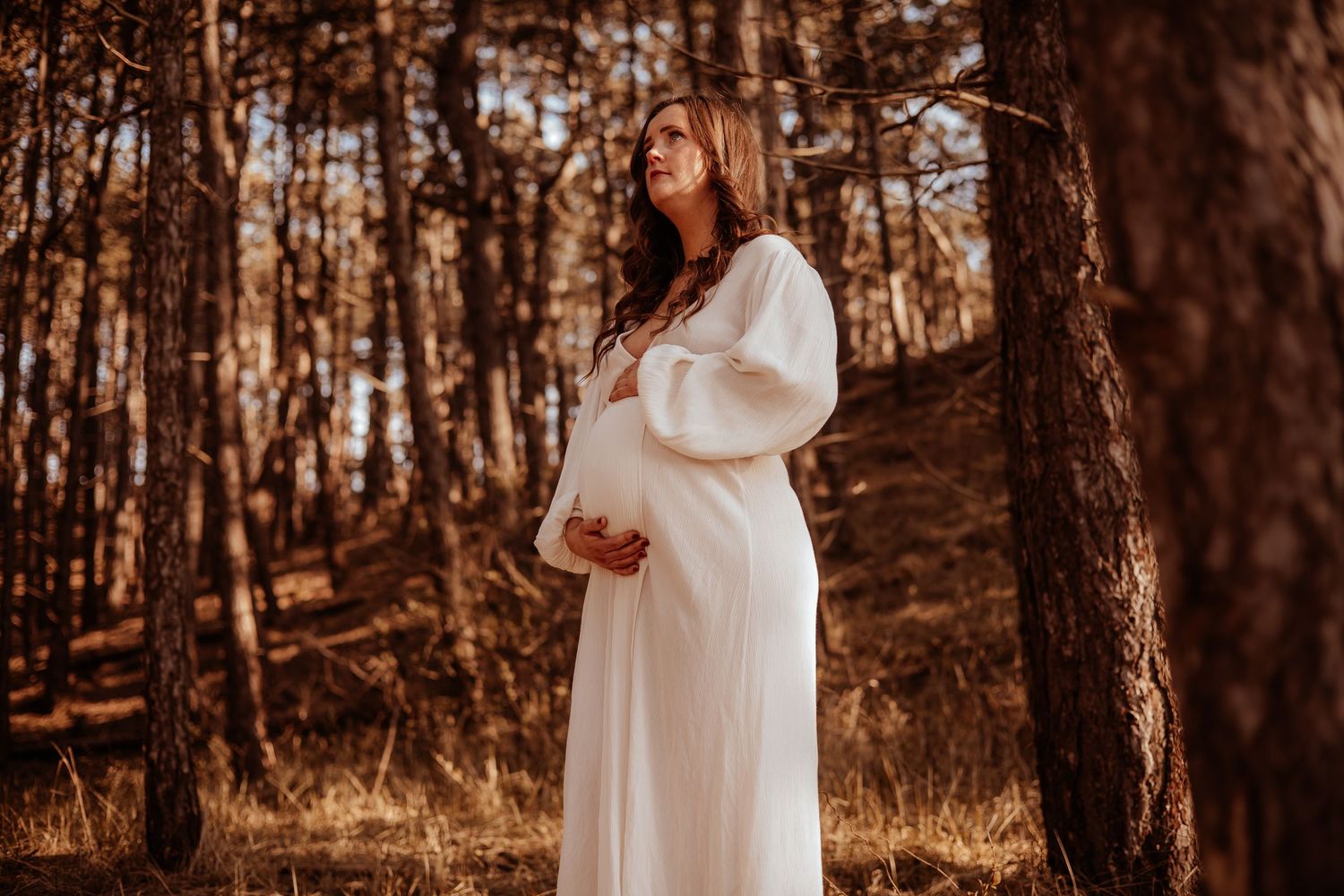 Pregnant woman in flowing white dress standing among pine trees, gazing upward in warm autumn light.