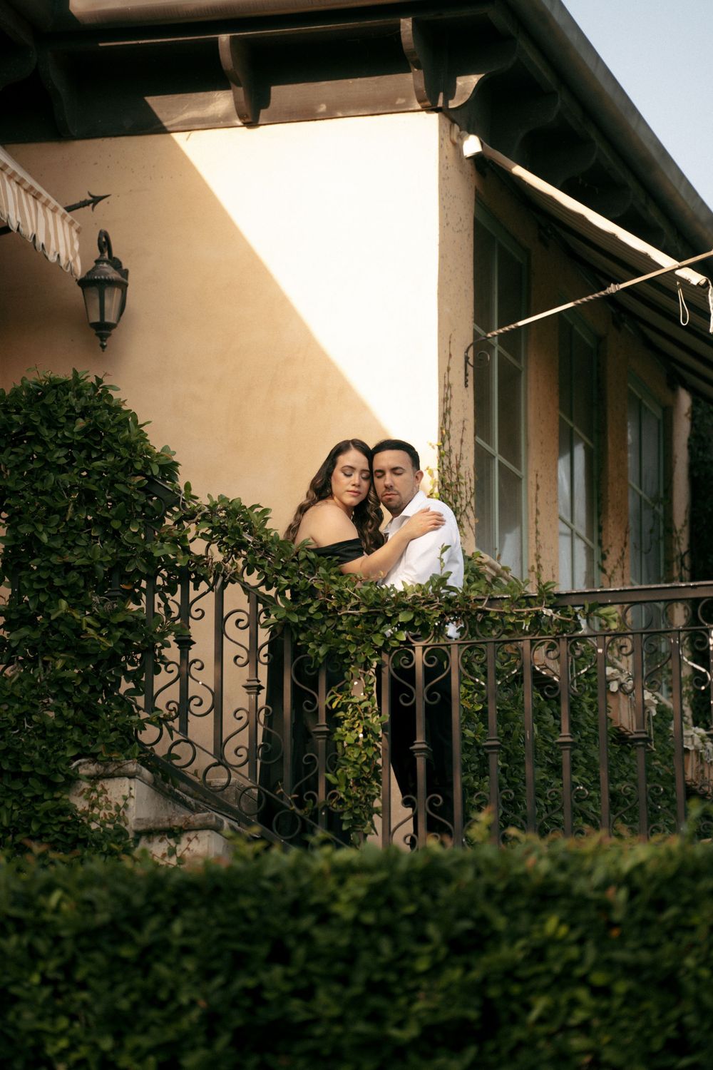 Couple embracing romantically on an ornate iron balcony surrounded by lush green hedges outside a Mediterranean-style home.