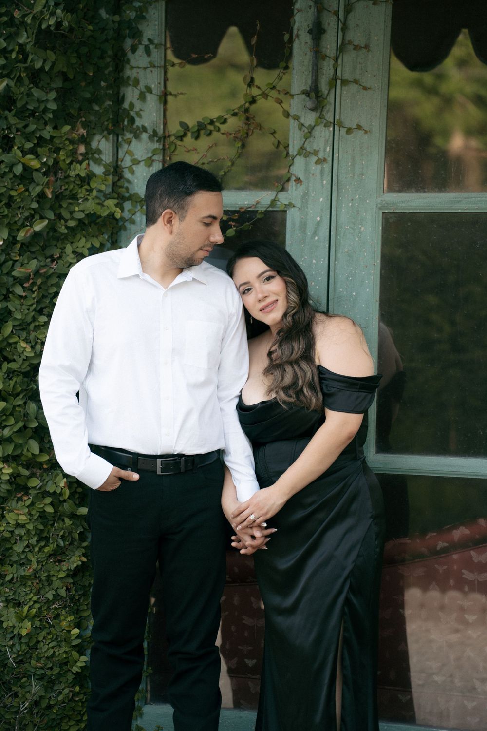 Couple in elegant black and white attire posing together near ivy-covered wall and vintage window outdoors.