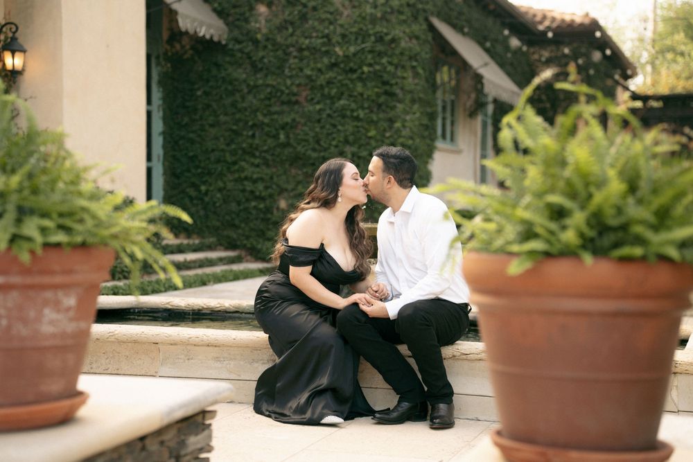Couple sharing a romantic kiss while sitting on stone steps surrounded by lush ivy and potted ferns at an elegant venue.