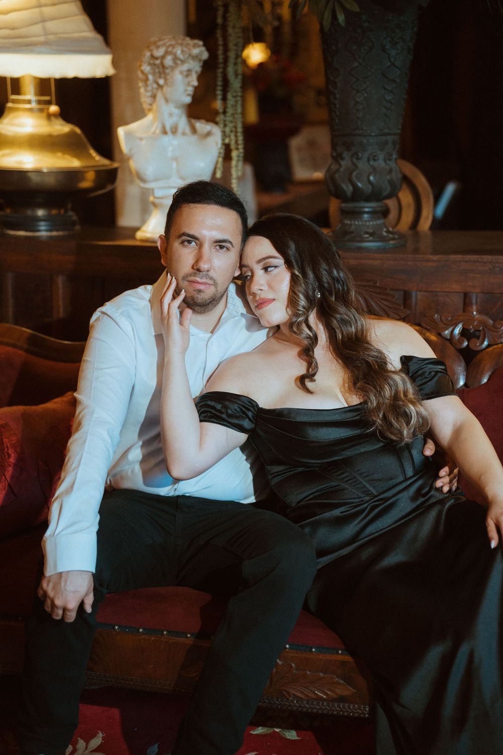 Elegant couple posing on ornate sofa, man in white shirt, woman in black off-shoulder gown, luxury vintage interior.