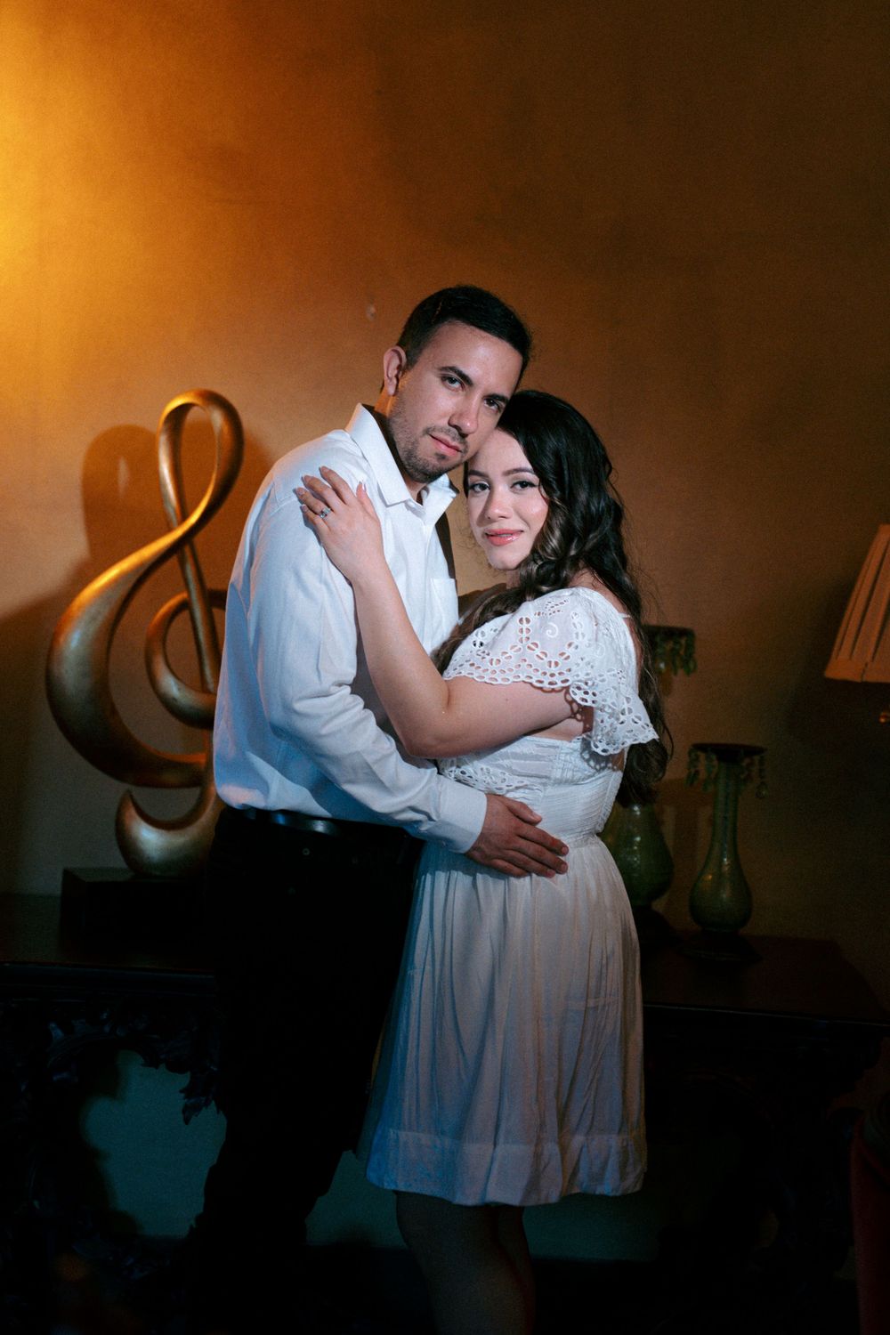 Romantic couple in white outfits embracing near a golden treble clef decoration in warm ambient lighting.