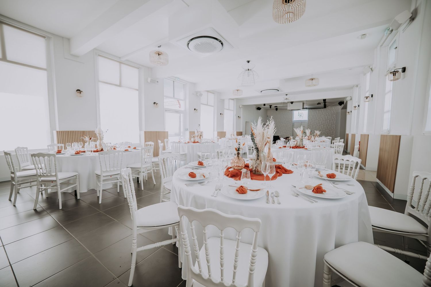 Elegant wedding reception hall with round tables, white tablecloths, red accents, and white chairs in a bright venue.