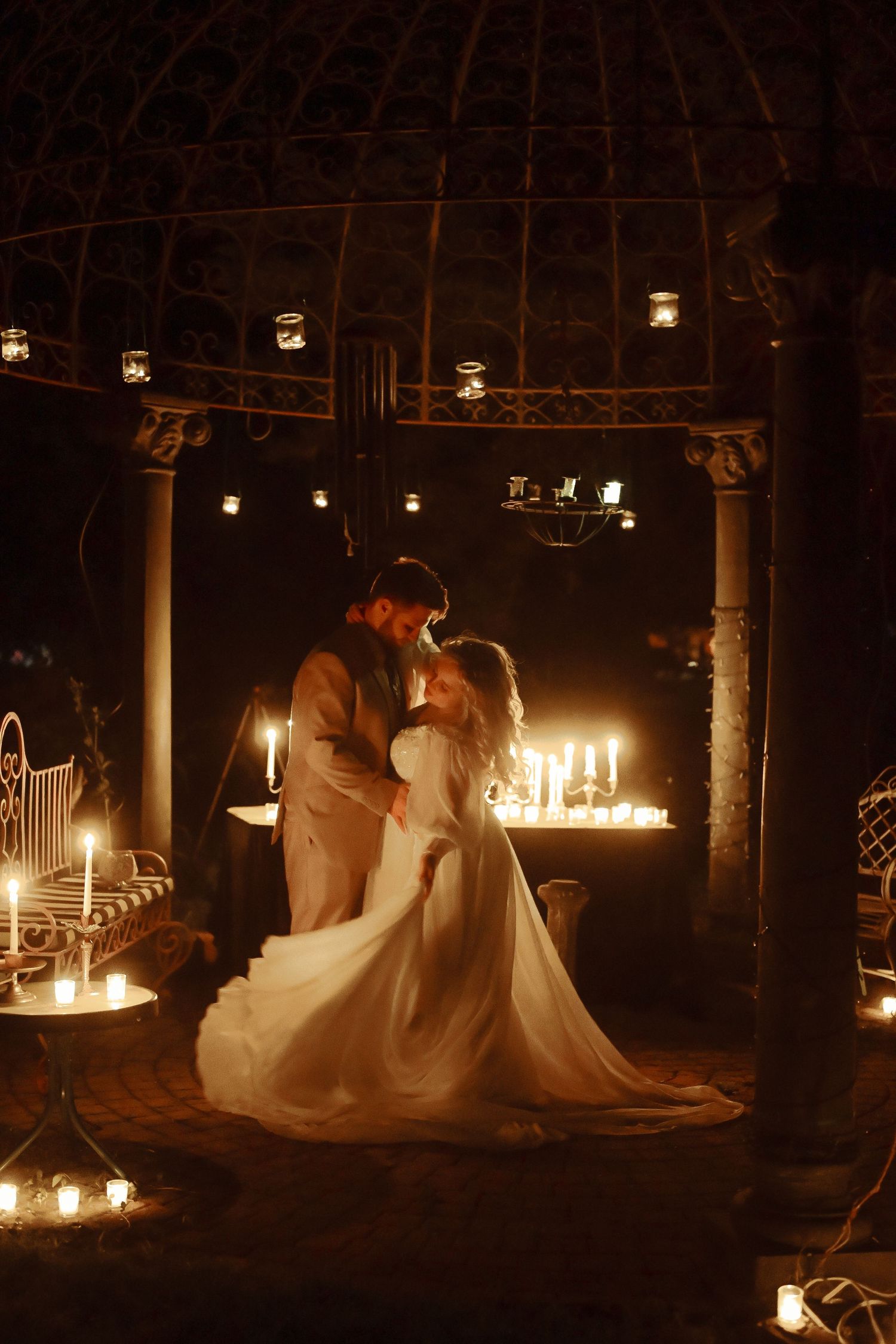Romantic bride and groom embracing by candlelight under an ornate gazebo during an intimate evening wedding ceremony.