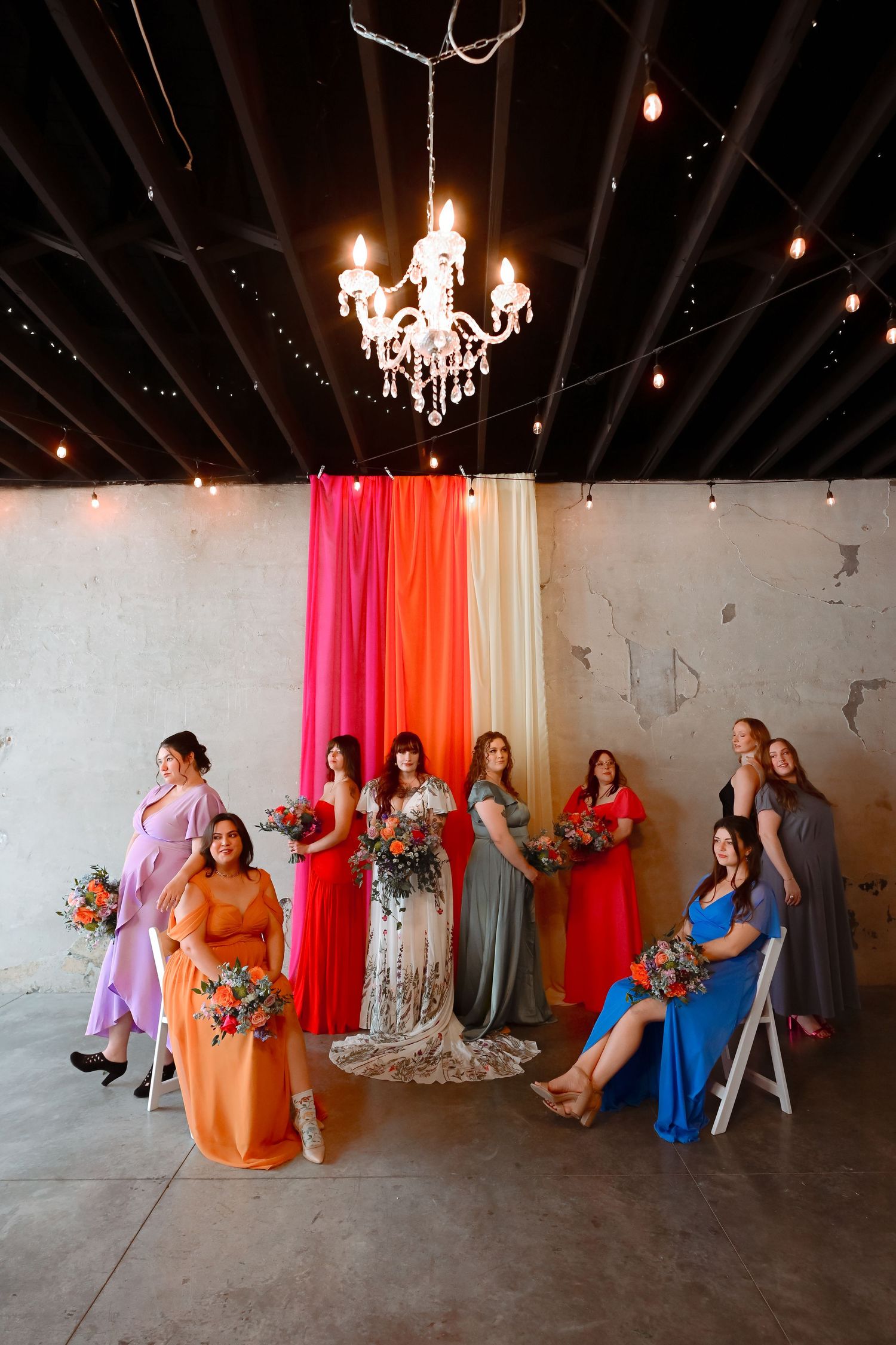Bride with colorful bridesmaids in mismatched dresses holding bouquets under chandelier with rainbow fabric backdrop.