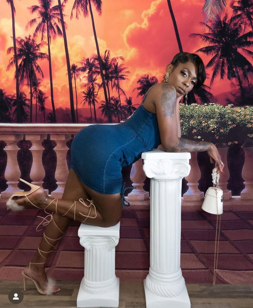 Woman in denim outfit posing on white columns with tropical sunset backdrop featuring palm trees.