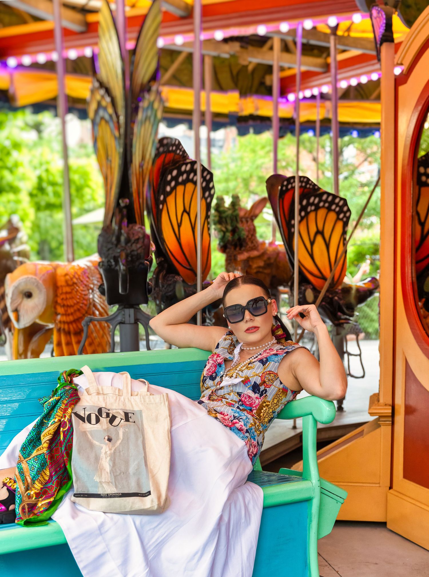 A stylish woman in sunglasses poses on a colorful carousel with monarch butterfly decorations and a Vogue tote bag.