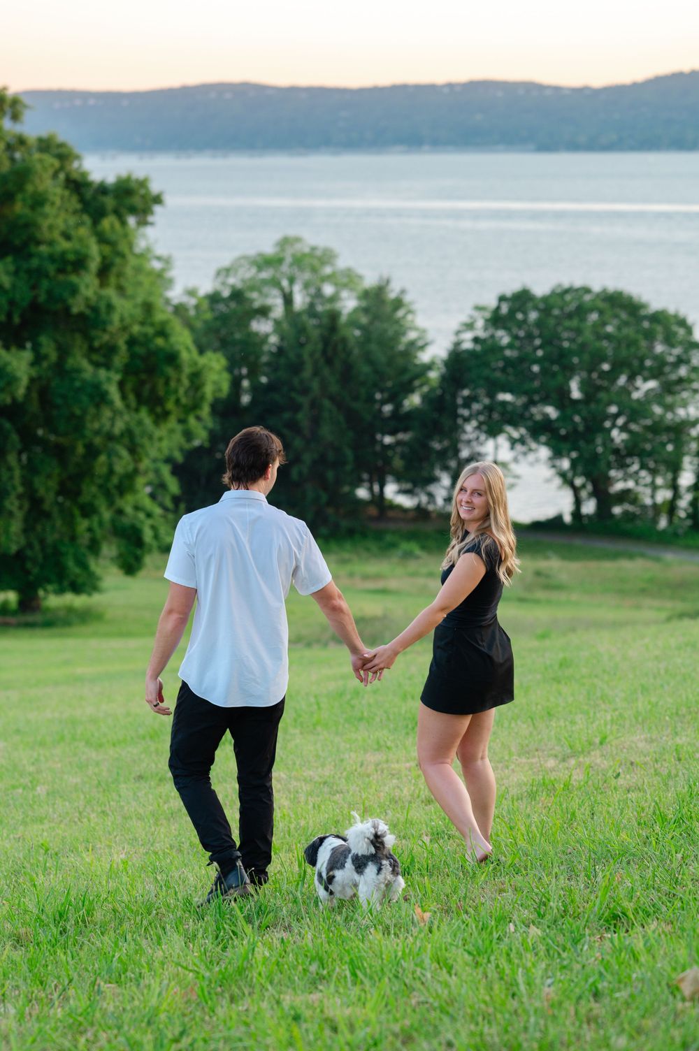 Couple holding hands on green hill with small dog, overlooking scenic lake and trees at sunset.