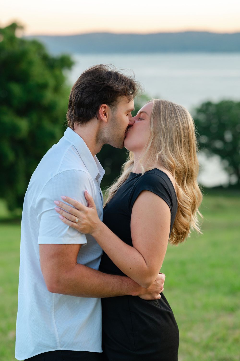 Couple sharing a romantic kiss outdoors at sunset with a scenic lake and green hills in the background.