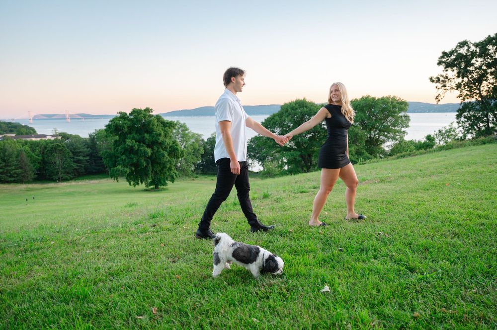 Happy couple holding hands and walking their Shih Tzu dog on a green hill at sunset by the water.