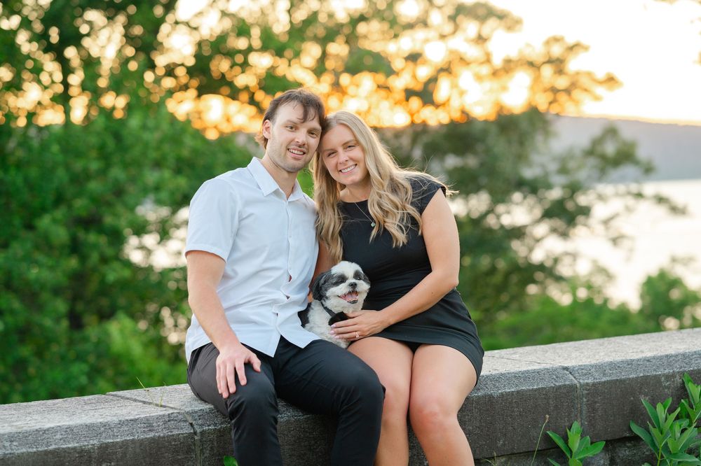 Smiling couple sitting on stone wall holding small dog at golden hour.