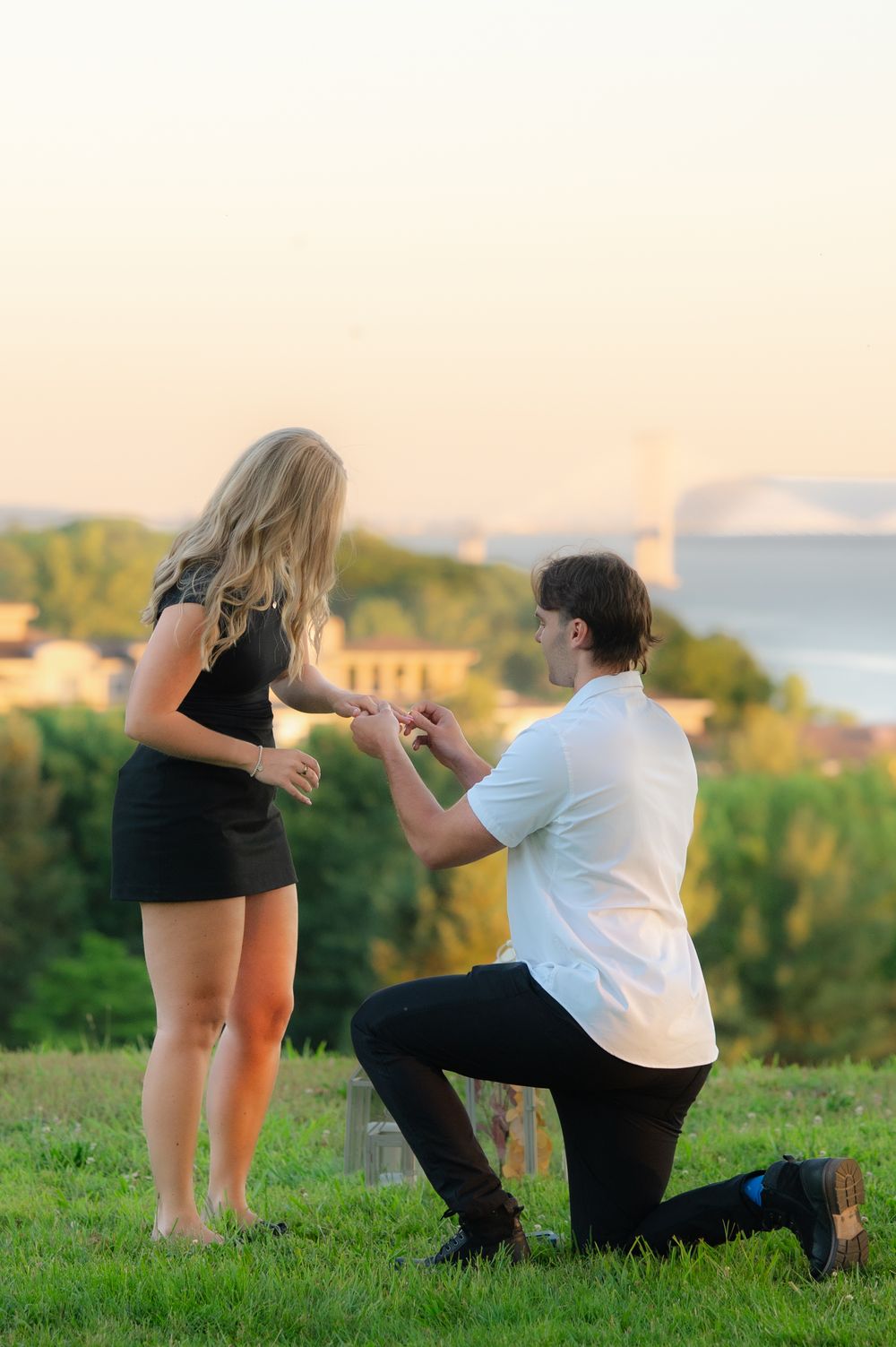 Man kneeling on one knee proposing to woman in black dress at sunset with scenic view in background.
