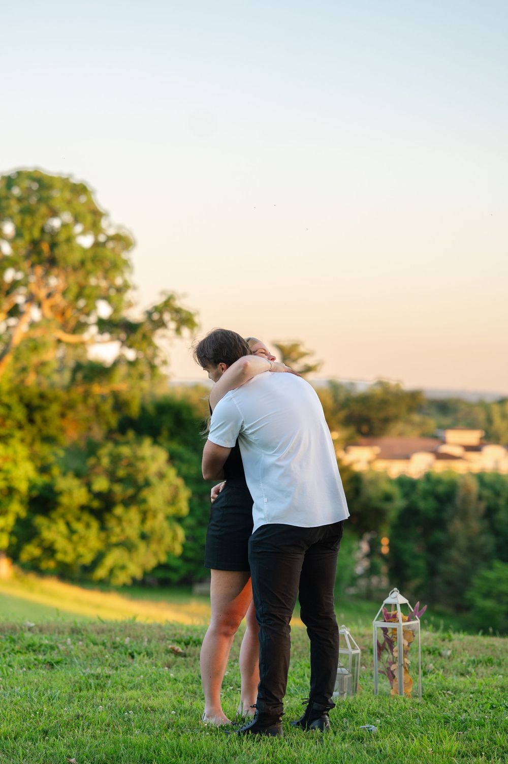 Couple embracing on a grassy hill at sunset with lanterns and lush green trees in the background.