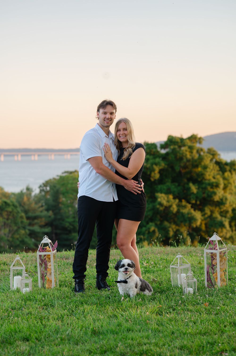 Smiling couple posing with their small Shih Tzu dog on a green hill at sunset with lanterns nearby.