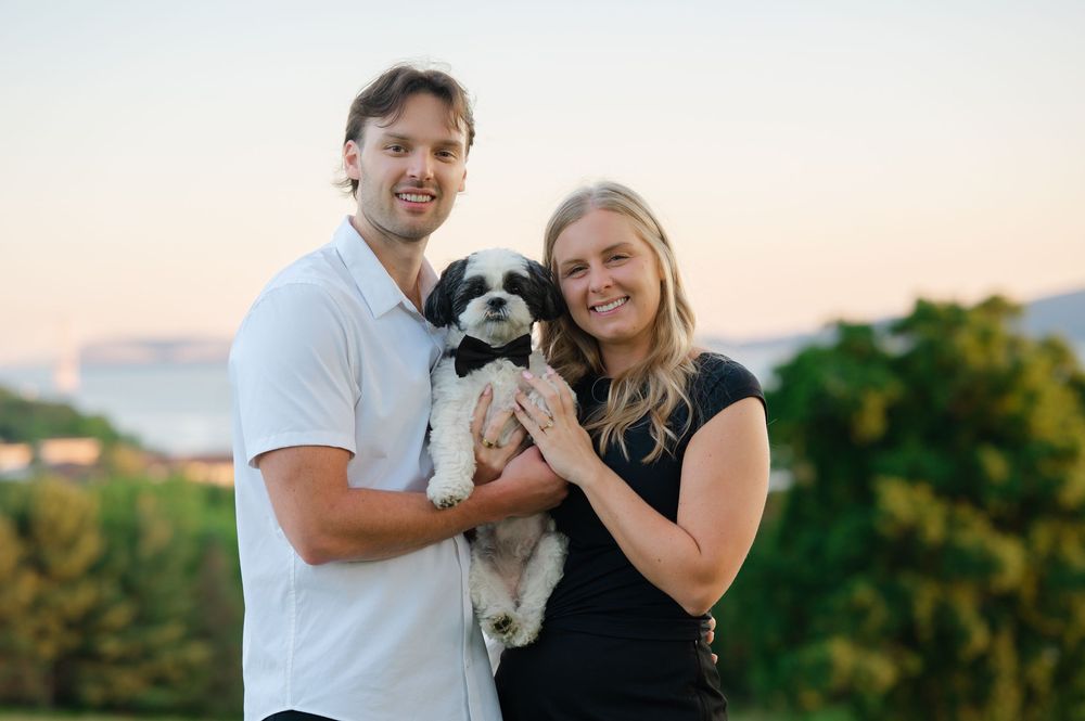 Smiling couple holding a black and white Shih Tzu dog outdoors at sunset with green hills in background.