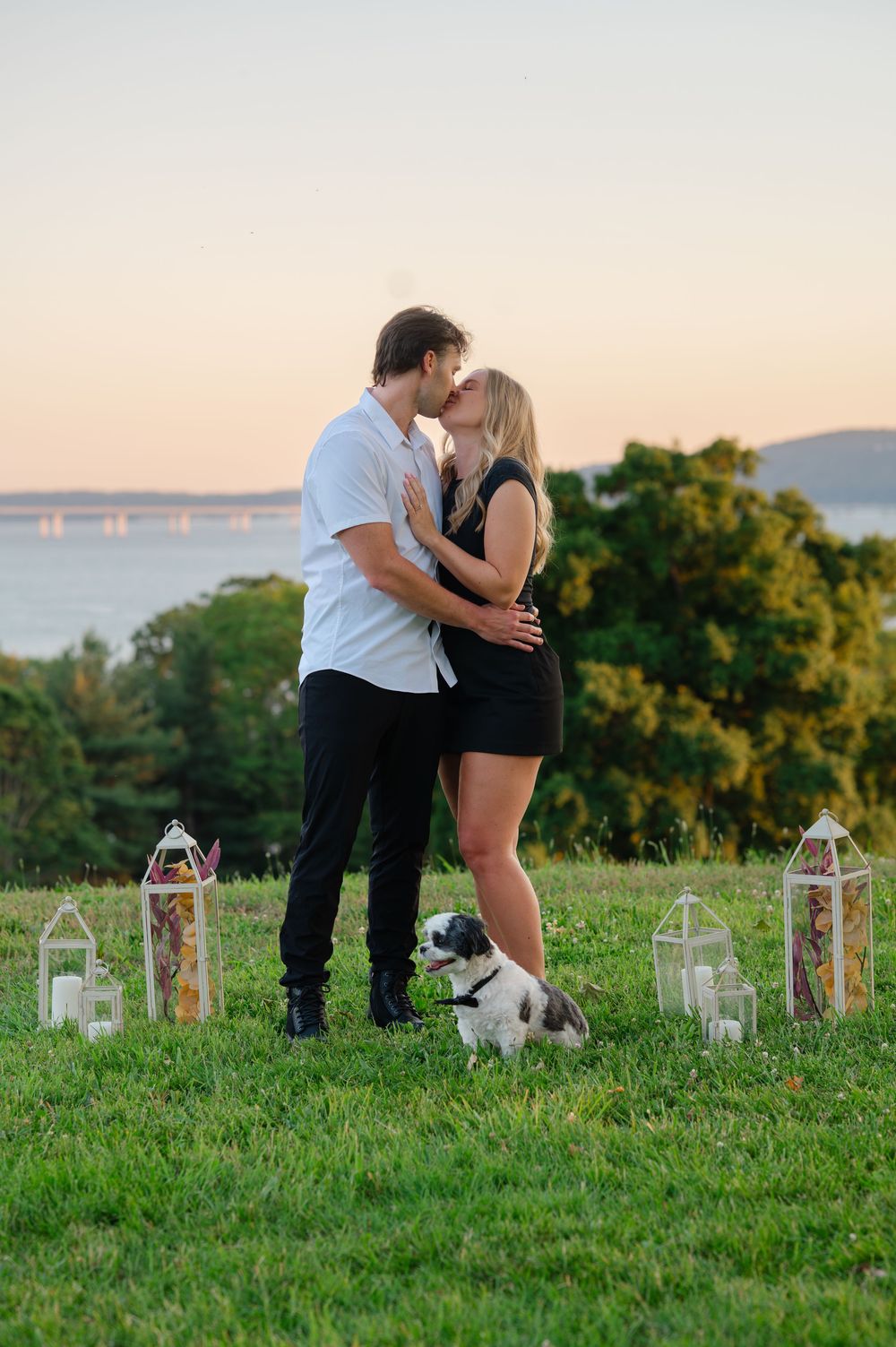 Couple kisses at sunset on grass with lanterns and a small dog at their feet near a scenic waterway.