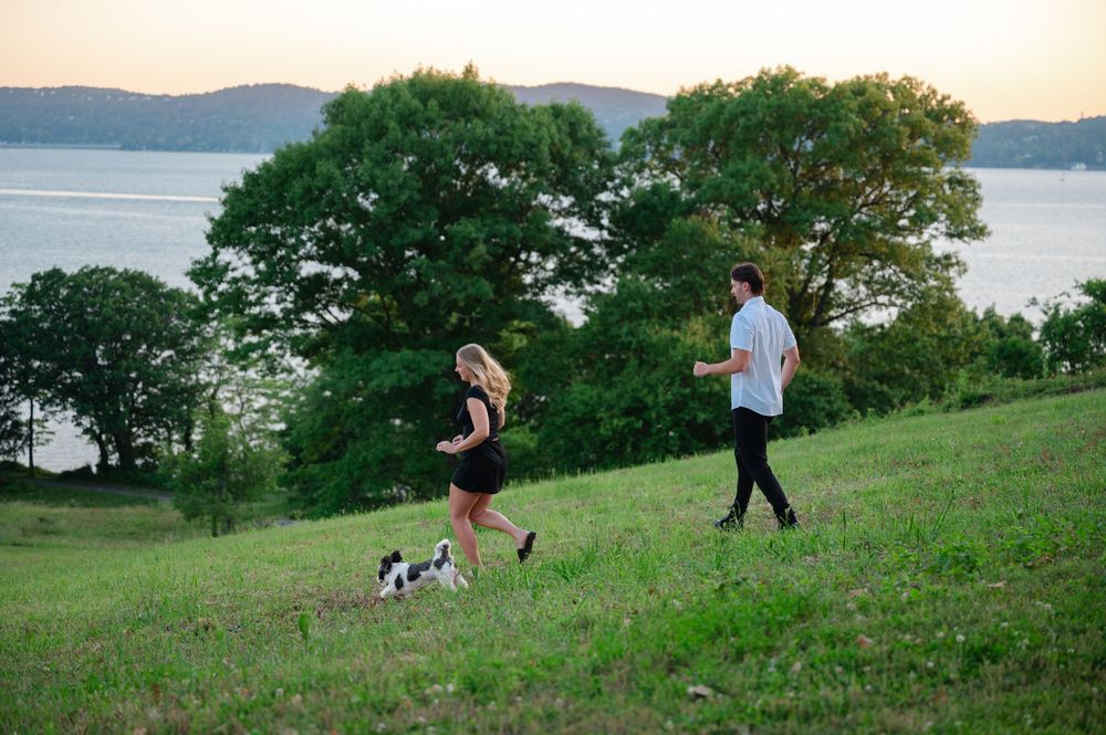 A couple jogging with a small dog on a green hillside overlooking a scenic lake at sunset.