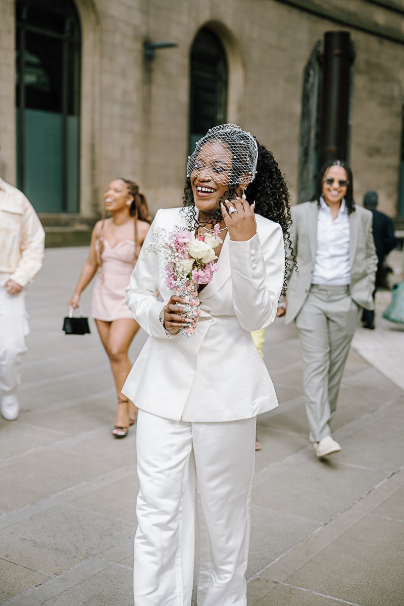 Joyful Black bride in white suit and birdcage veil holds bouquet while talking on phone outdoors.