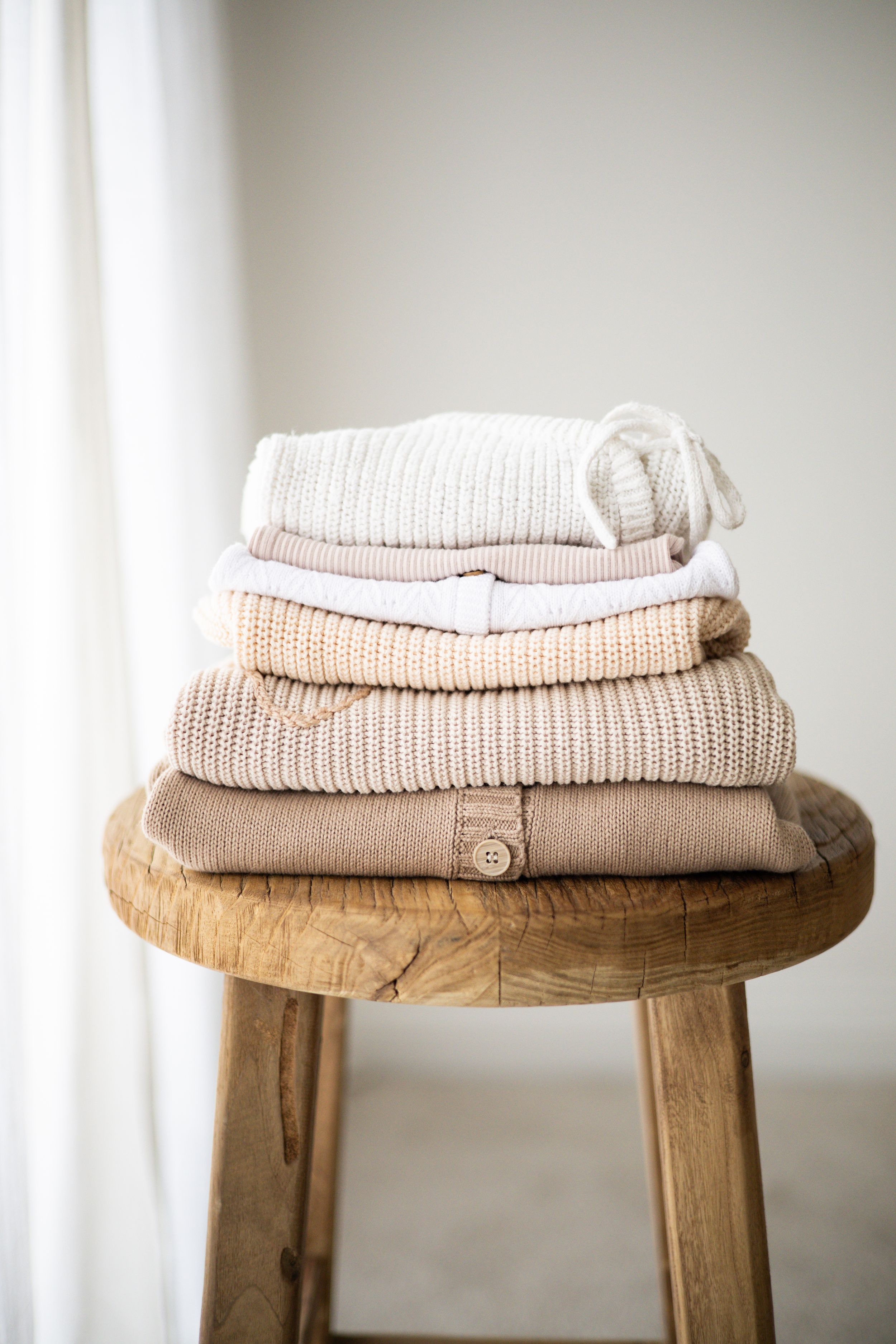 Stack of folded knit sweaters in neutral tones on a rustic wooden stool near a bright window Newborn photographer Nottingham