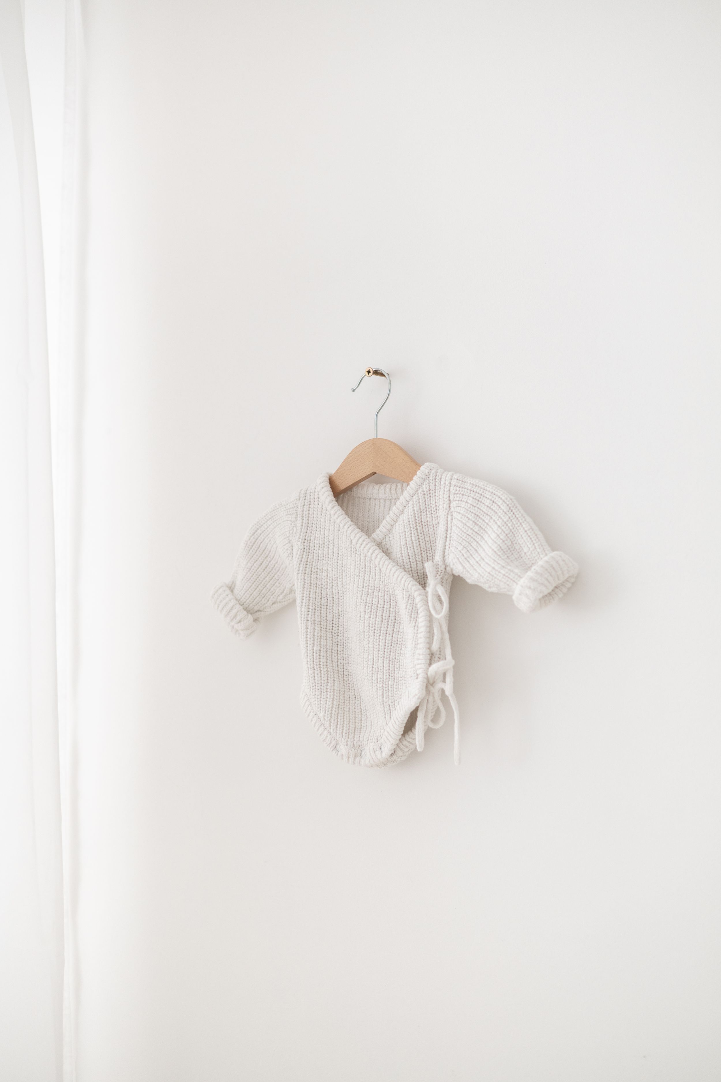 Tiny white knit wrap bodysuit for newborns hanging on a wooden hanger against a white wall Newborn photographer Nottingham