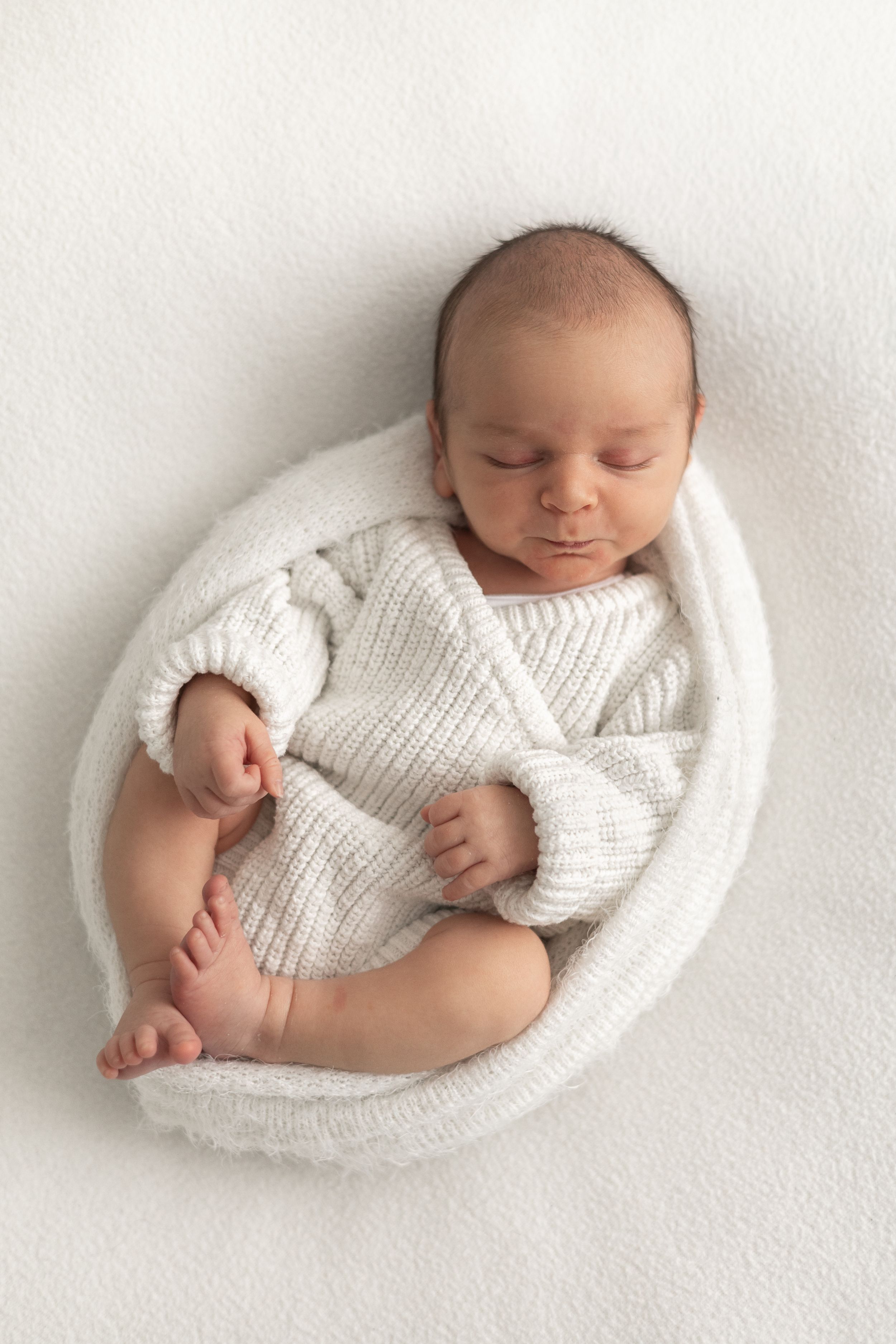 Peaceful newborn baby sleeping curled up in white knit wrap and outfit on soft white background Newborn photographer Nottingham
