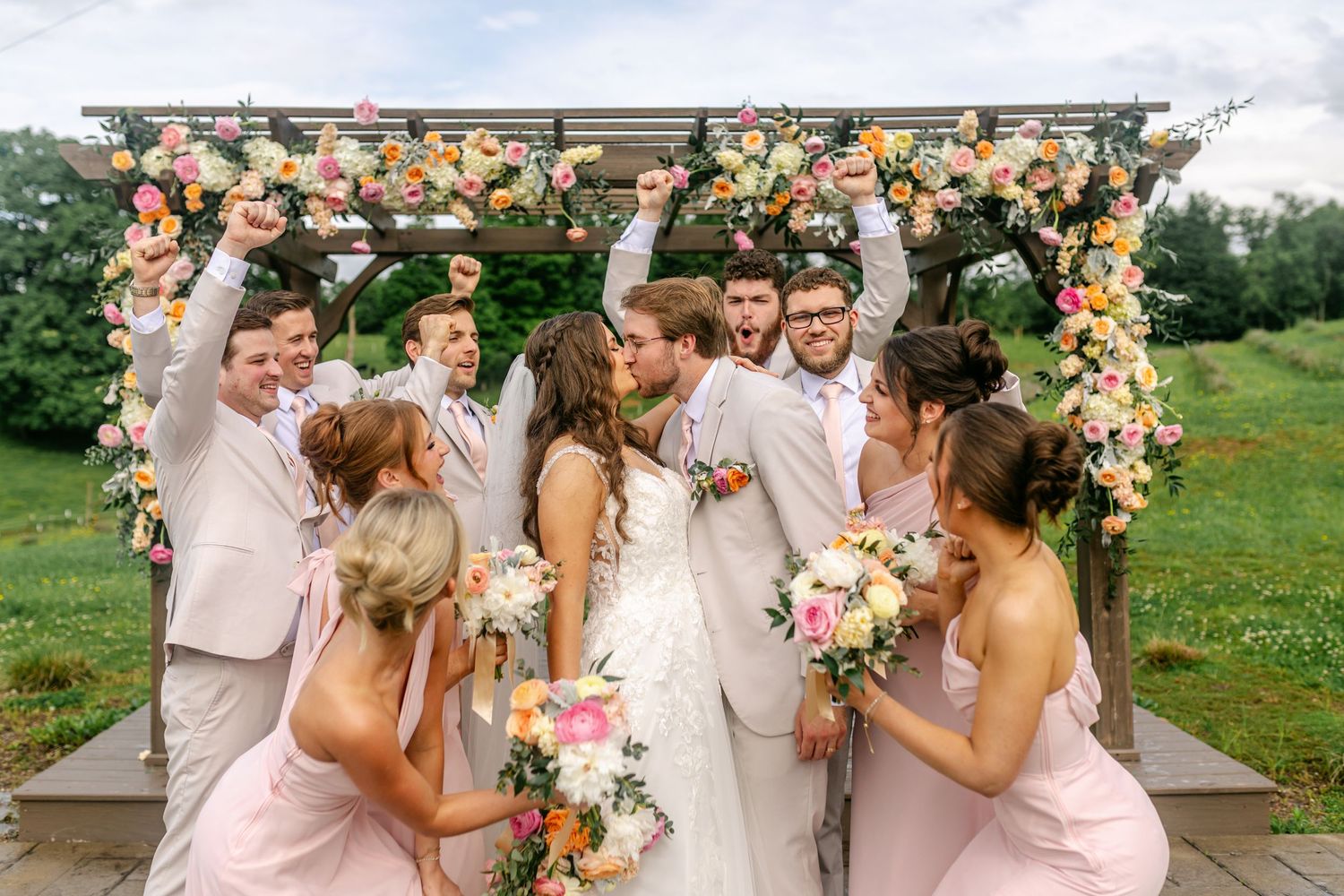 Excited wedding party surrounds kissing bride and groom under floral pergola with colorful spring blooms.