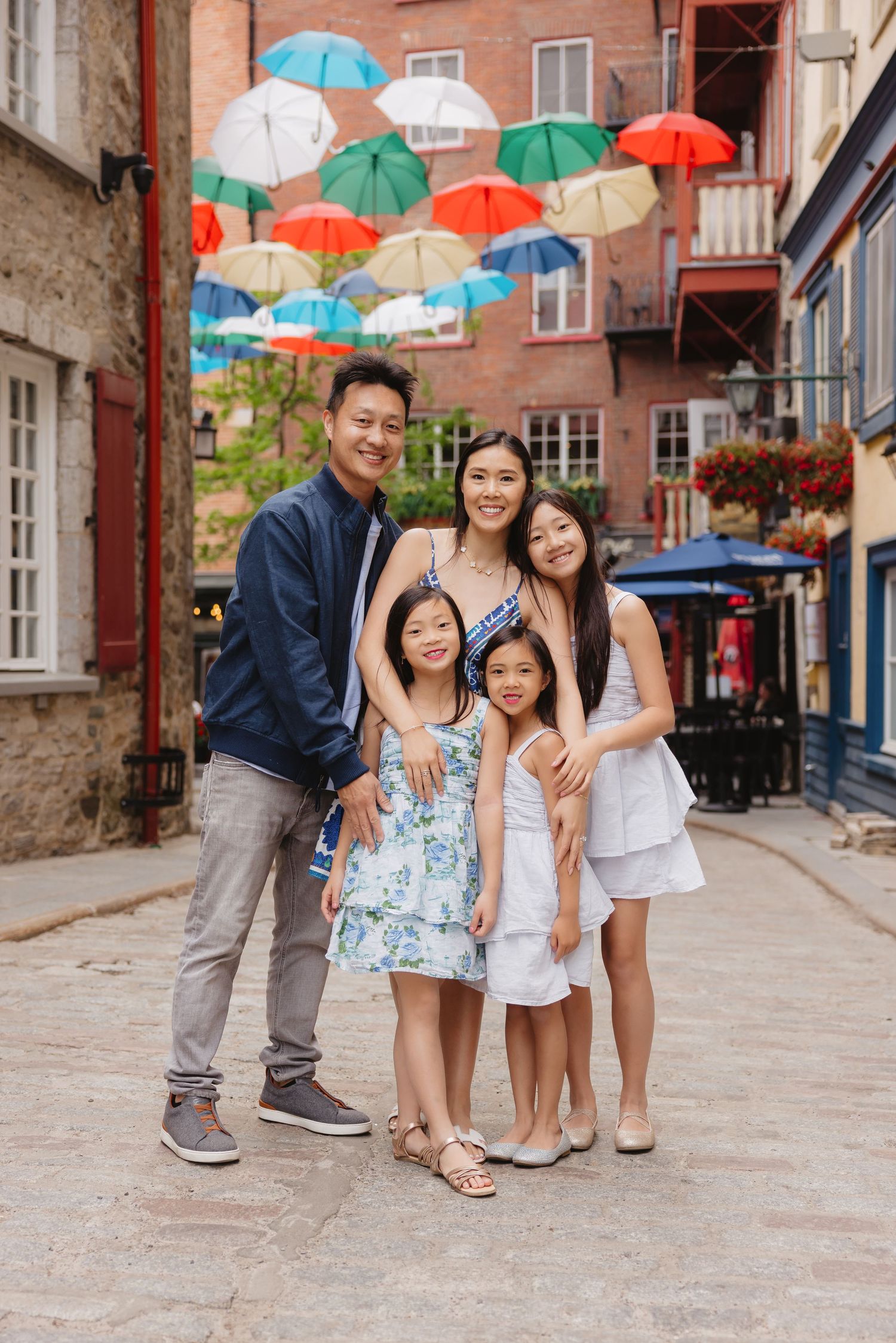 Asian family of five posing on cobblestone street with colorful floating umbrellas overhead in historic Quebec City.
