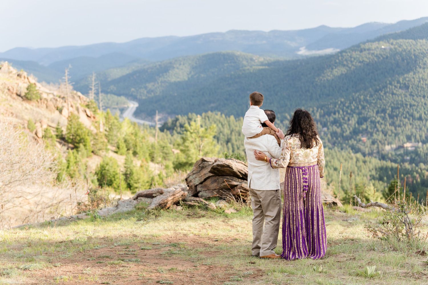 Family of three standing on a mountain overlook, admiring lush green forested valley views in Colorado.