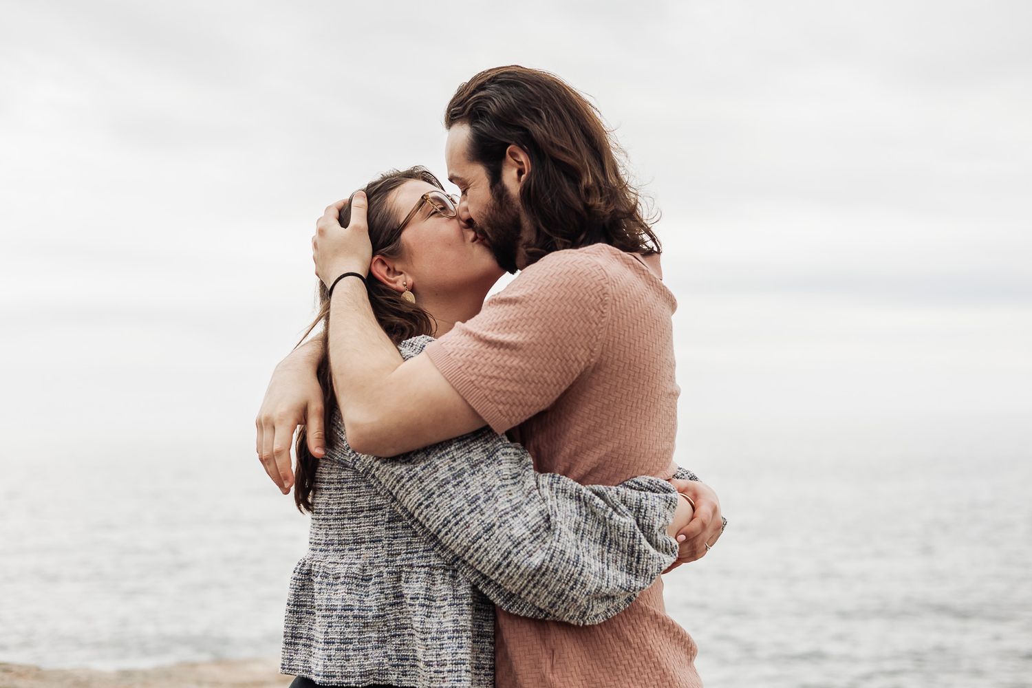 Romantic couple embracing and kissing by the ocean on a cloudy day, woman in gray jacket and man with long hair.