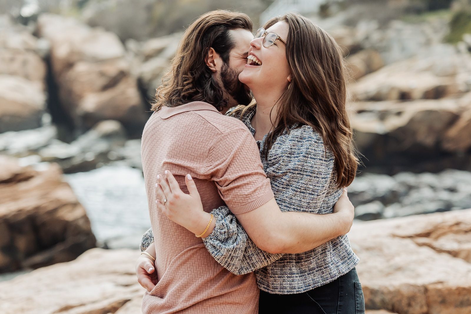 A joyful couple embracing and laughing together on a rocky beach, sharing a romantic outdoor moment.