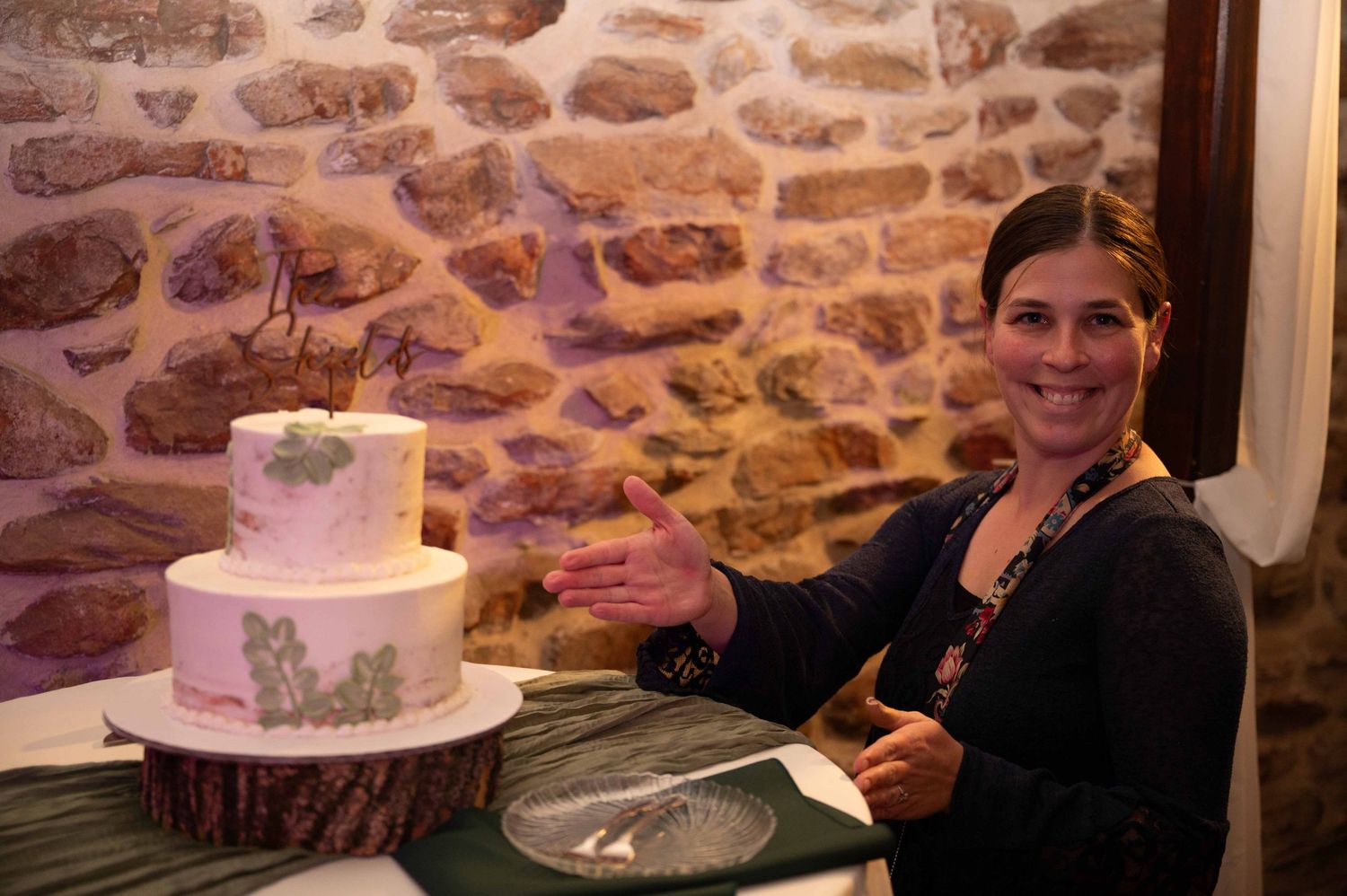 A server stands next to an elegant two-tier white cake with floral designs against a rustic stone wall.