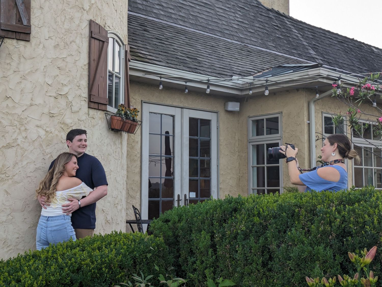 People stand outside a residential home with stucco walls, French doors, and neatly trimmed hedges in the front.