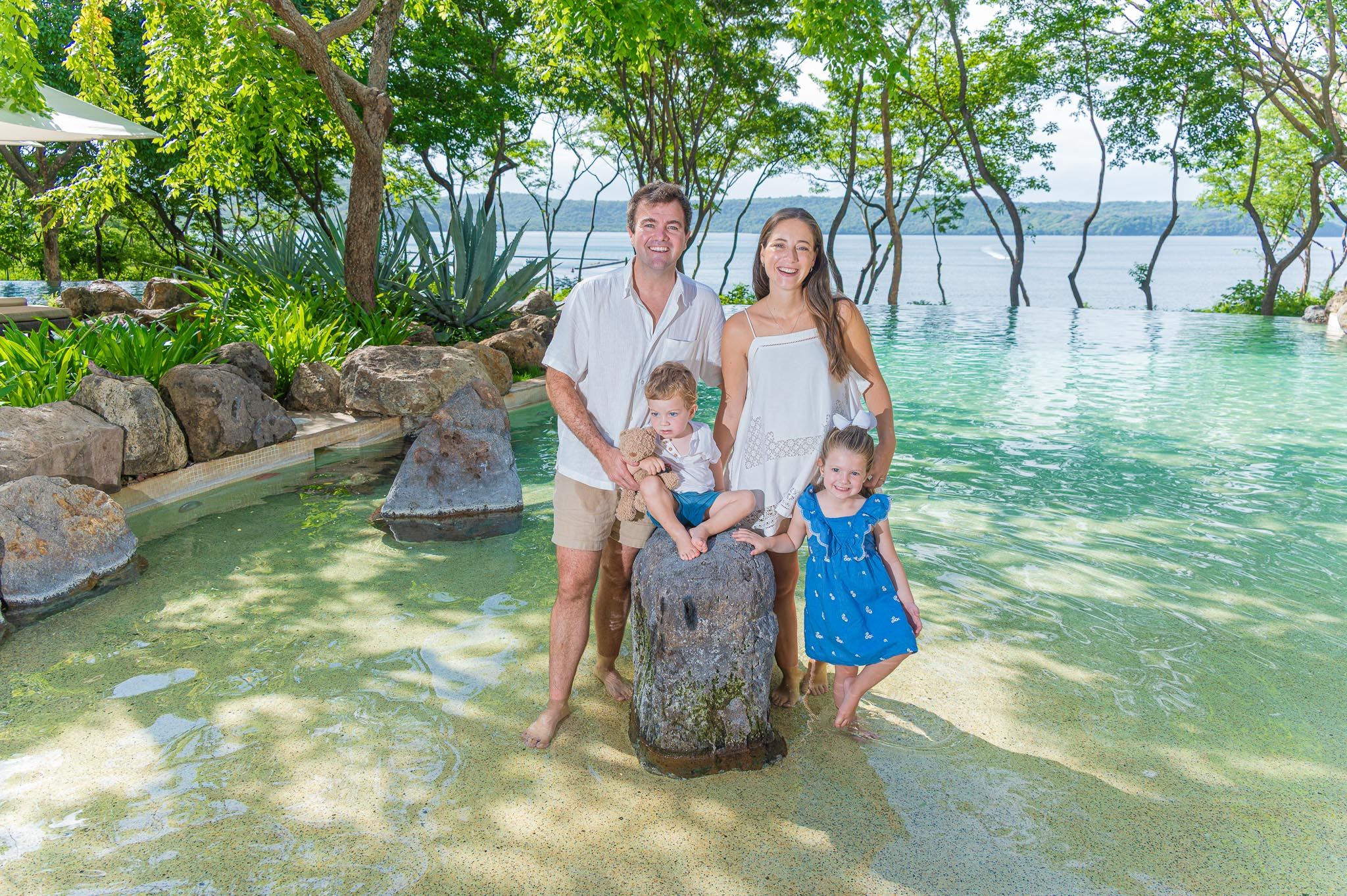 Family of four poses by a tropical pool with lush greenery and calm water in a resort setting.