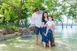 Family of three enjoying shallow tropical waters near lush green trees and rocky shore on a sunny day.