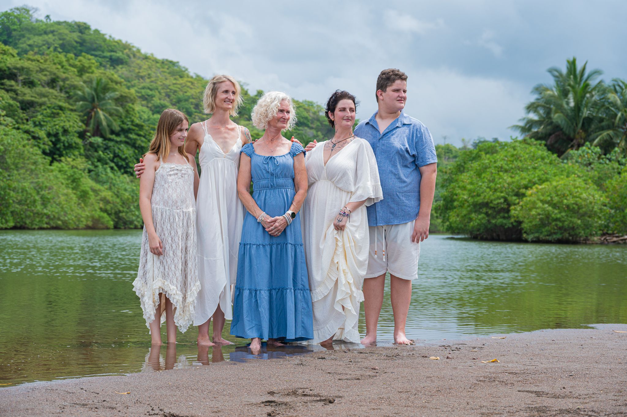 Family of five posing on a tropical beach shore with lush green mangroves in the background.
