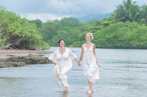 Two brides in white dresses hold hands walking through shallow water on a tropical beach.