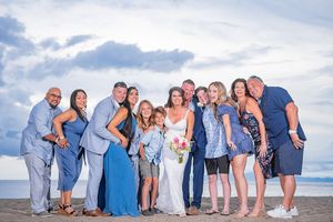 Wedding party posing together on a beach, wearing blue outfits, with the bride holding a bouquet under a cloudy sky.