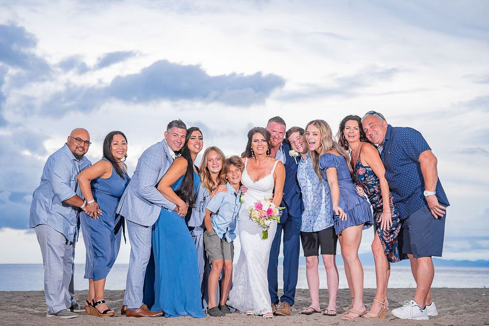 Wedding party posing together on a beach, wearing blue outfits, with the bride holding a bouquet under a cloudy sky.