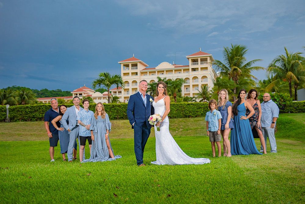 Wedding couple poses with bridal party on lush lawn before tropical resort under dramatic cloudy sky.